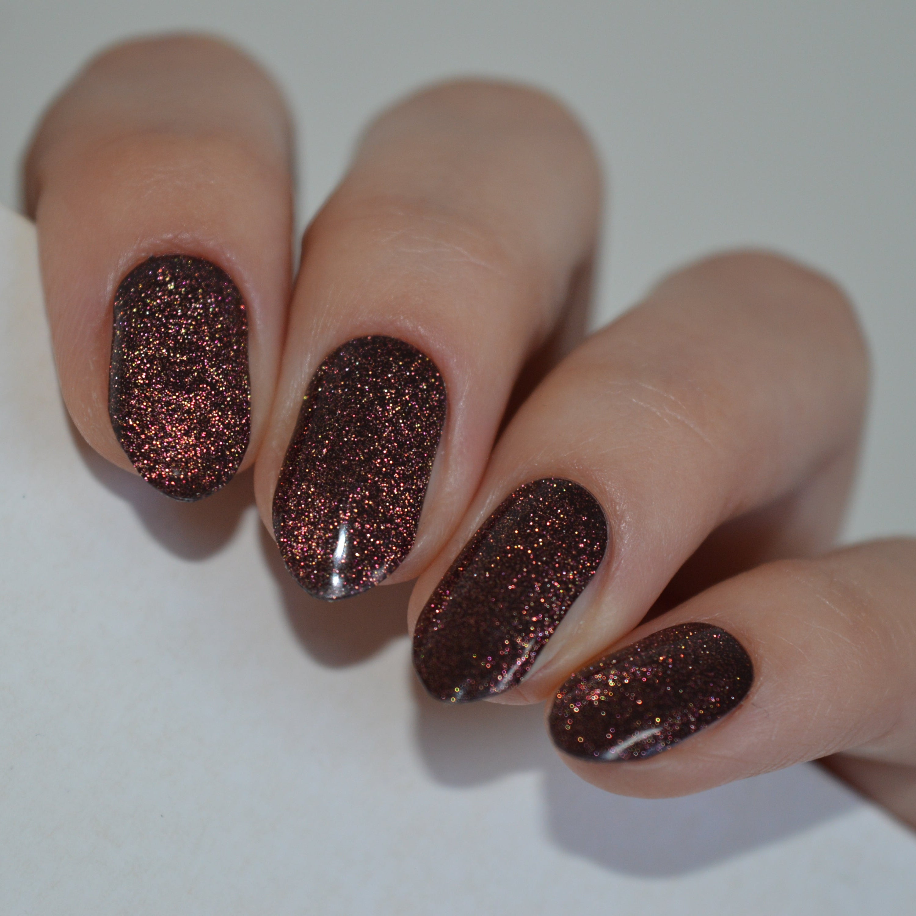 A close-up of a hand with manicured nails painted in a dark brown glitter nail polish, resting on a plain white surface.