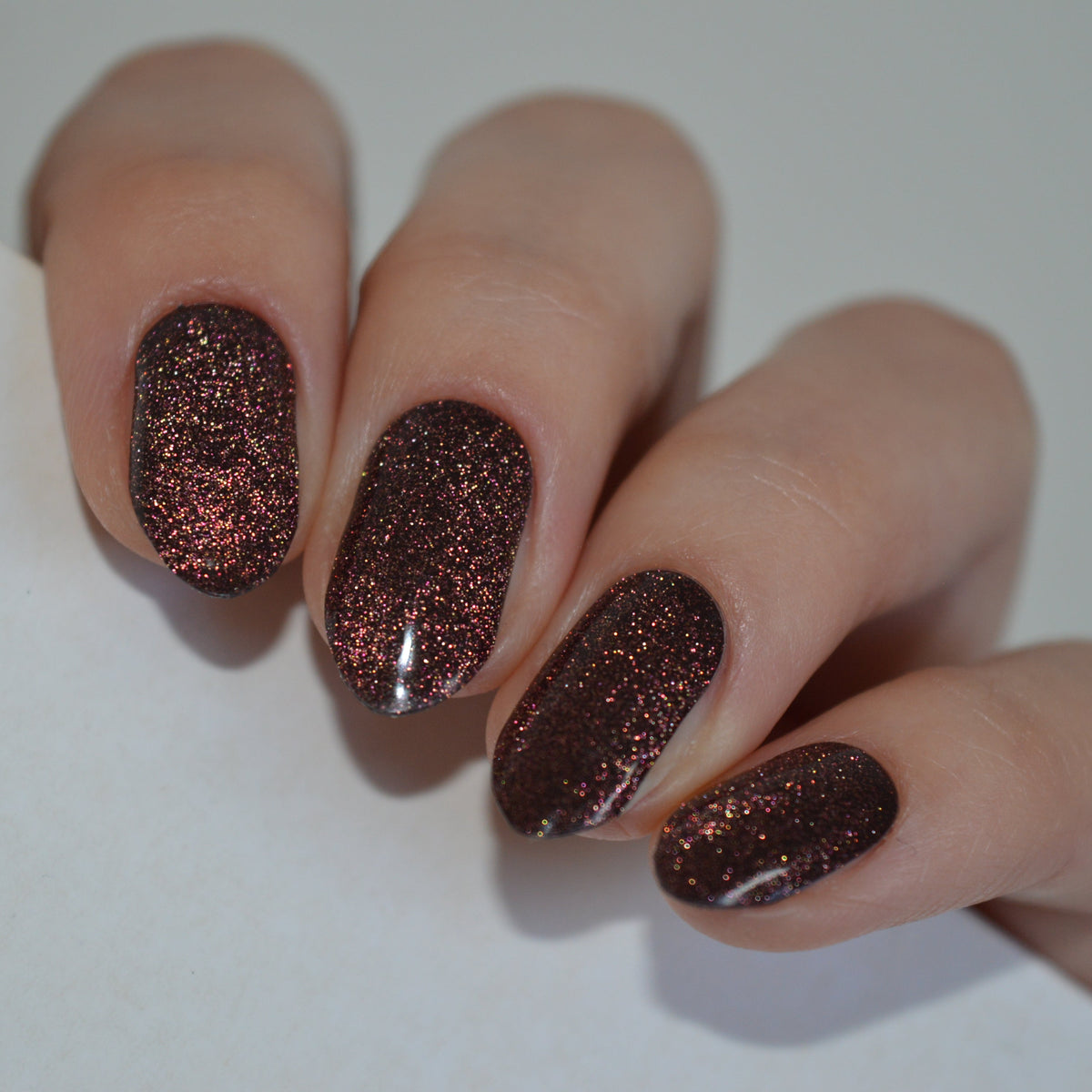 A close-up of a hand with manicured nails painted in a dark brown glitter nail polish, resting on a plain white surface.